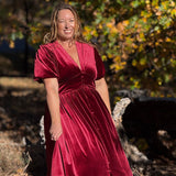 A woman wearing a deep burgundy velvet midi dress with short puff sleeves and a V-neckline, standing outdoors in front of a blurred background of autumn trees.