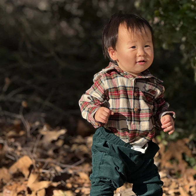 A toddler standing outdoors on a log surrounded by dry leaves and trees, wearing a plaid button-up shirt with dark green pants.