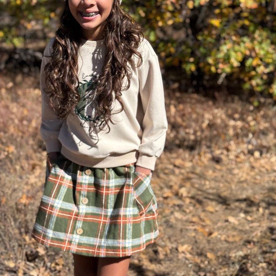 A young girl smiling outdoors in a cream knit beanie, a cream sweatshirt, and a green plaid button-up skirt with orange and white stripes.
