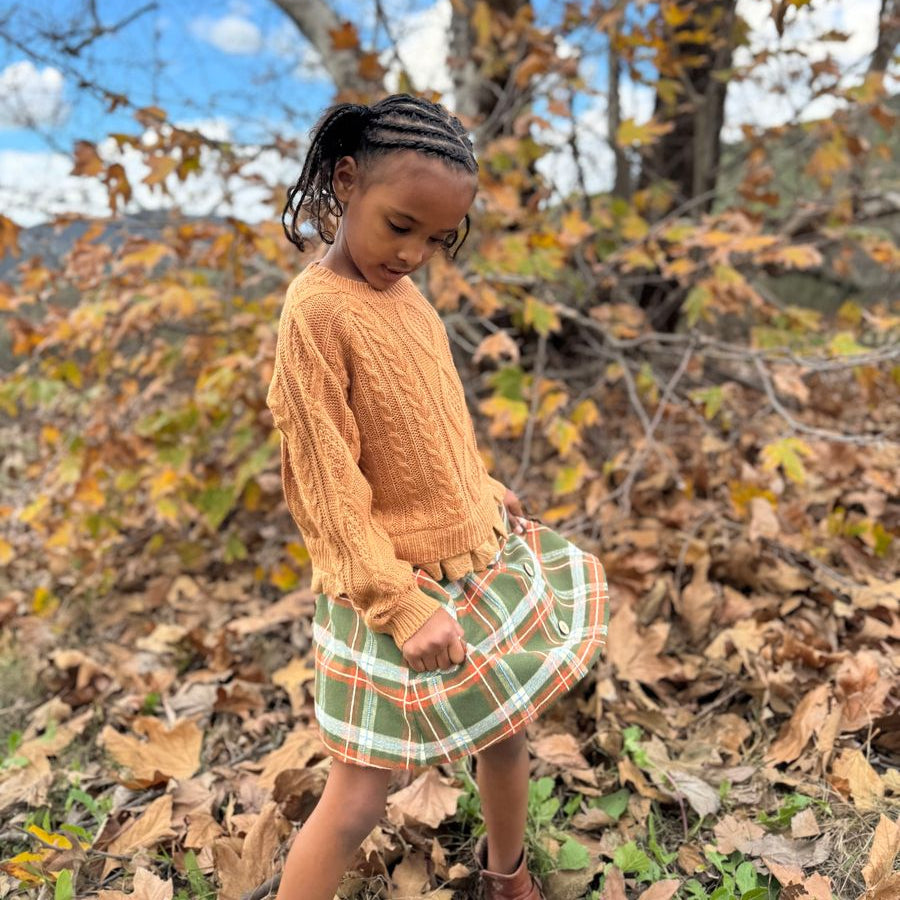 A young girl wearing a textured mustard yellow cable-knit sweater and a green, orange, and white plaid skirt, standing outdoors in a wooded area covered with fallen autumn leaves.