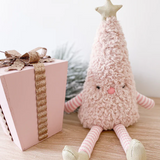 A soft plush Christmas tree doll with a fuzzy pink body, striped pink and white arms and legs, a beige star on its head, and a small pink face, posed on a wooden surface beside a pink gift box with a gold ribbon.