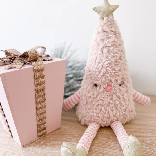 A soft plush Christmas tree doll with a fuzzy pink body, striped pink and white arms and legs, a beige star on its head, and a small pink face, posed on a wooden surface beside a pink gift box with a gold ribbon.
