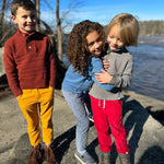 Three children standing outdoors by a river under a clear blue sky, wearing cozy knit sweaters and pants in red, yellow, blue, gray, and pink.
