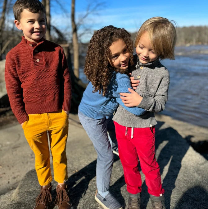 Three children standing outdoors by a river under a clear blue sky, wearing cozy knit sweaters and pants in red, yellow, blue, gray, and pink.