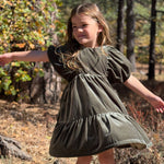 A young girl with long light brown hair stands outdoors in a wooded area wearing an olive green tiered dress with puff sleeves. The background features trees, dry leaves, and natural sunlight.