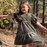 A young girl with long light brown hair stands outdoors in a wooded area wearing an olive green tiered dress with puff sleeves. The background features trees, dry leaves, and natural sunlight.