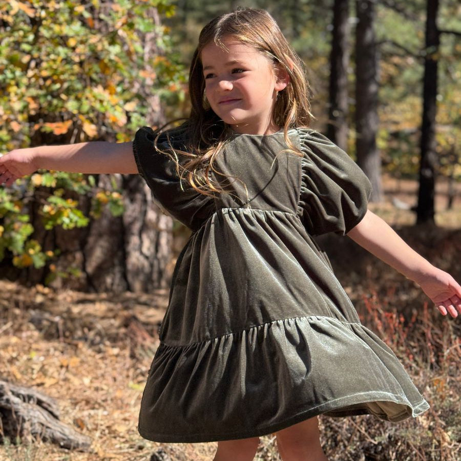 A young girl with long light brown hair stands outdoors in a wooded area wearing an olive green tiered dress with puff sleeves. The background features trees, dry leaves, and natural sunlight.