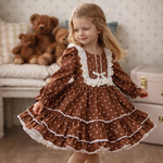 A young girl with blonde hair wearing a brown and white polka dot dress with white lace trim, standing in a cozy room with teddy bears and a wooden floor.
