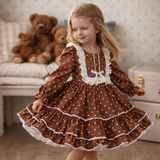 A young girl with blonde hair wearing a brown and white polka dot dress with white lace trim, standing in a cozy room with teddy bears and a wooden floor.