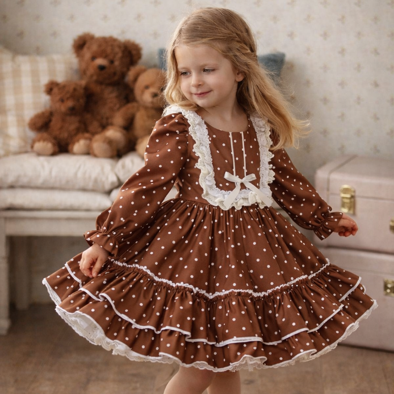 A young girl with blonde hair wearing a brown and white polka dot dress with white lace trim, standing in a cozy room with teddy bears and a wooden floor.