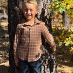 A young boy wearing a brown and red plaid button-down shirt and blue jeans stands outdoors leaning against a large tree trunk in a wooded area with autumn foliage.