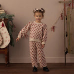 A young girl stands in a cozy room wearing a cream-colored two-piece pajama set with a red floral pattern. The set includes a long-sleeve top and matching pants.