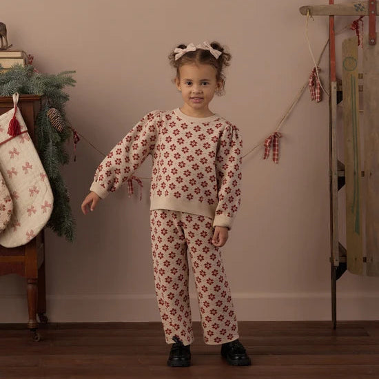 A young girl stands in a cozy room wearing a cream-colored two-piece pajama set with a red floral pattern. The set includes a long-sleeve top and matching pants.