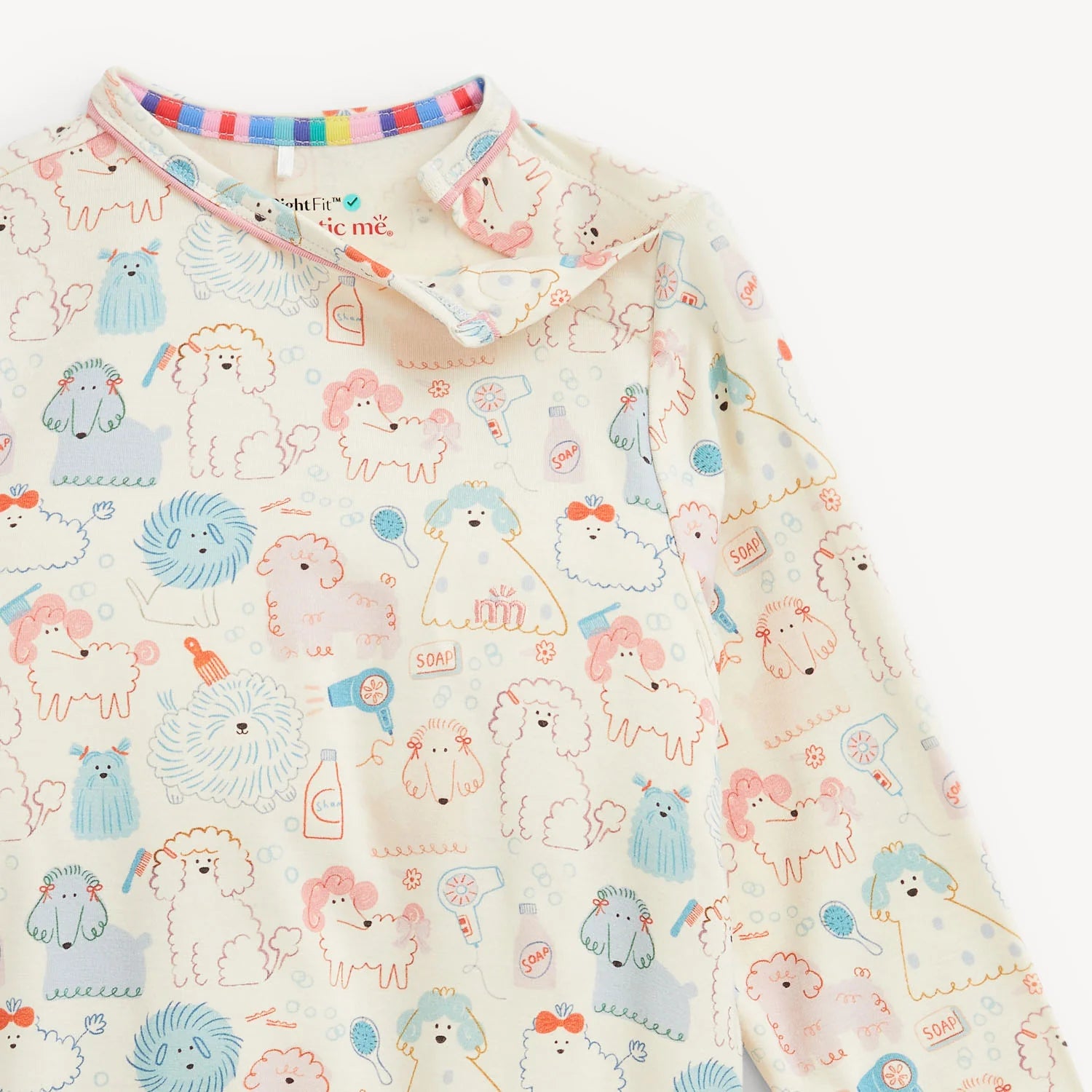 A close-up of a long-sleeve baby top featuring a cute cartoon dog and grooming-themed pattern in pastel colors on an off-white base. The collar has a colorful striped inner lining.