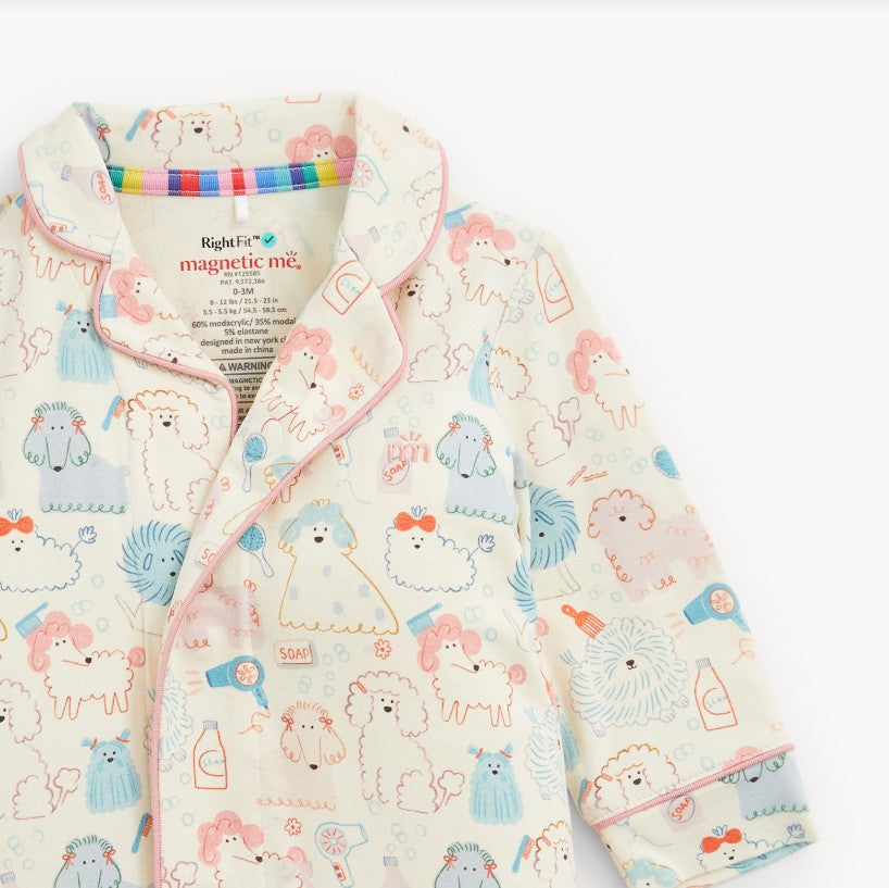 A close-up of a children's pajama top with a cute cartoon dog and grooming accessory print on a cream background, featuring pink piping and a visible inner neck label reading 'magnetic me'.