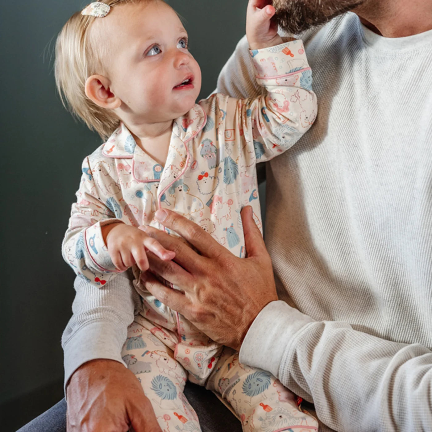 A baby wearing a long-sleeve pajama romper with a colorful animal print sits on an adult's lap. The pajamas feature a light cream base with blue, pink, and orange cartoon animals.