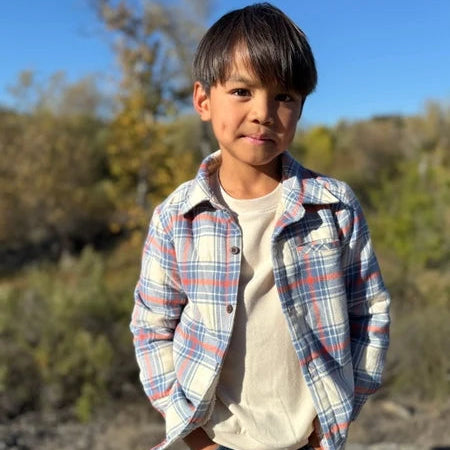 A young boy wearing a blue, red, and white plaid button-down shirt over a cream t-shirt and dark jeans, standing outdoors in a sunny, natural setting with trees and rocks in the background.