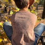 A child seen from behind wearing a brown cowboy hat, a faded tan graphic tank top with 'SHOULDA BEEN A COWBOY' and blue jeans, sitting outdoors in front of autumn trees.