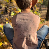 A child seen from behind wearing a brown cowboy hat, a faded tan graphic tank top with 'SHOULDA BEEN A COWBOY' and blue jeans, sitting outdoors in front of autumn trees.