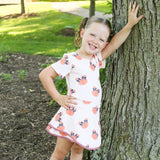 A young girl smiling and posing outdoors against a tree trunk, wearing a 2 piece athletic set with a pink and orange pumpkin print and brown lace-up shoes.