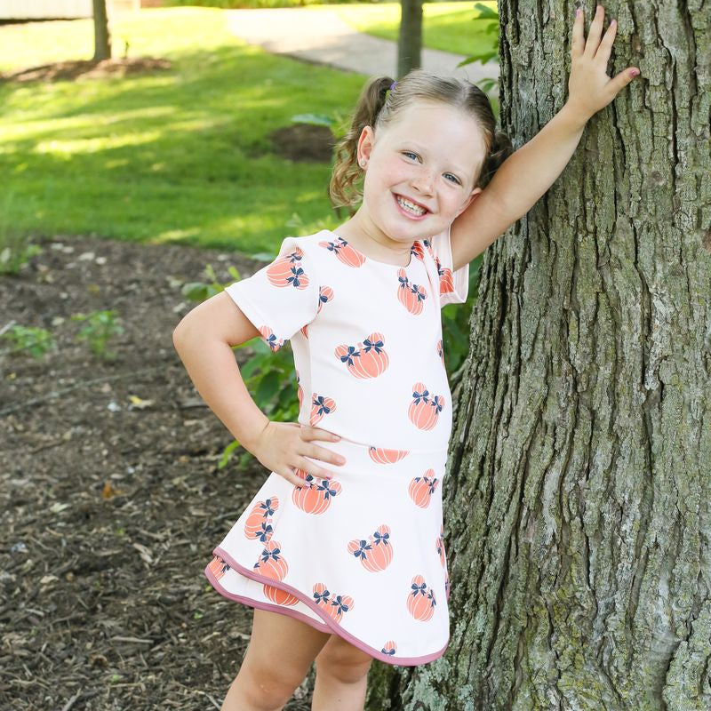 A young girl smiling and posing outdoors against a tree trunk, wearing a 2 piece athletic set with a pink and orange pumpkin print and brown lace-up shoes.