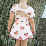 A young girl viewed from behind wearing a short-sleeve cream 2 piece set with an orange pumpkin print and a pink back cutout, standing outdoors in front of a large tree trunk.