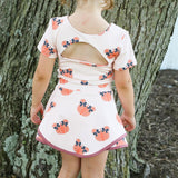A young girl viewed from behind wearing a short-sleeve cream 2 piece set with an orange pumpkin print and a pink back cutout, standing outdoors in front of a large tree trunk.