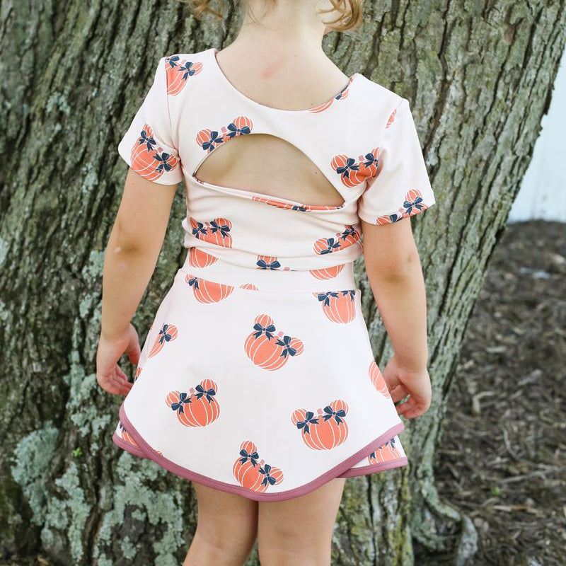 A young girl viewed from behind wearing a short-sleeve cream 2 piece set with an orange pumpkin print and a pink back cutout, standing outdoors in front of a large tree trunk.