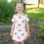 A young girl with blonde pigtails stands outdoors on a mulch path wearing a  short-sleeve 2 piece set with a pattern of orange pumpkins and blue bows, paired with brown lace-up boots.