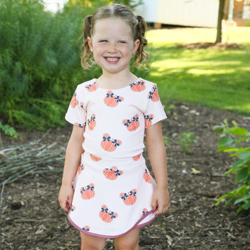 A young girl with blonde pigtails stands outdoors on a mulch path wearing a  short-sleeve 2 piece set with a pattern of orange pumpkins and blue bows, paired with brown lace-up boots.