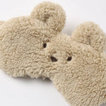 A close-up of a plush teddy bear face mask or face mask in a soft beige sherpa fabric, featuring embroidered dark brown eyes and nose on a clean white background.