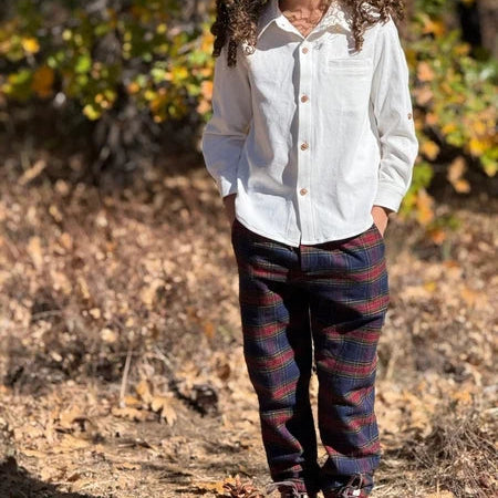 Child wearing a white shirt and plaid pants standing in an outdoor setting with trees and fallen leaves.