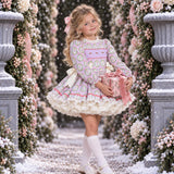 A young girl stands in a snowy garden archway wearing a long-sleeve ruffled dress with a pastel multicolor pattern, holding a pink gift box. The scene is framed by stone pillars and floral garlands.