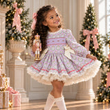 A young girl with curly brown hair wearing a lavender and white patterned long-sleeve dress with a ruffled skirt, white lace socks, and gold shoes, holding a nutcracker in a decorated holiday room with Christmas trees and columns.