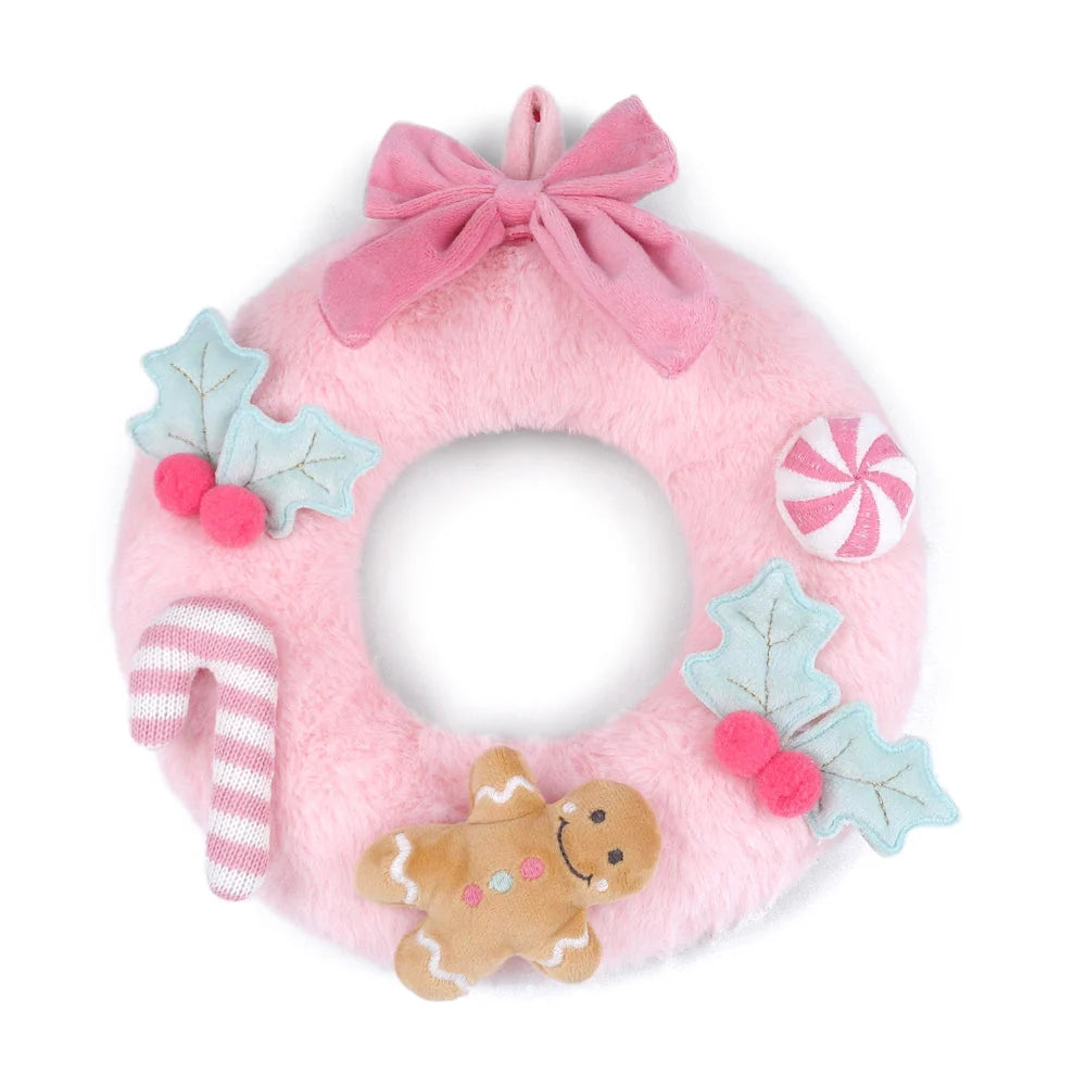 A pink plush felt Christmas wreath with a large pink bow at the top, decorated with light blue holly leaves with pink berries, a pink and white striped candy cane, a pink and white striped peppermint candy, and a brown gingerbread cookie ornament, all on a white background.