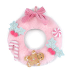 A pink plush felt Christmas wreath with a large pink bow at the top, decorated with light blue holly leaves with pink berries, a pink and white striped candy cane, a pink and white striped peppermint candy, and a brown gingerbread cookie ornament, all on a white background.